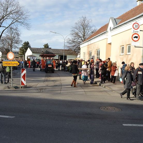 Eine Menschenmenge versammelt sich auf einer Straße, einige gehen, während andere in der Nähe eines Gebäudes stehen, an dem ein Schild mit der Aufschrift 'Umleitung' angebracht ist. Ein Traktor fährt auf der Straße.