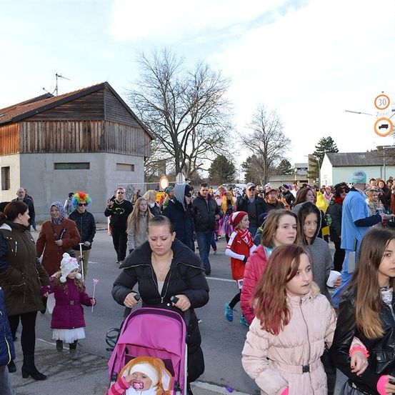 Eine Menschenmenge, darunter Kinder in Kostümen, geht auf einer Straße mit Gebäuden und Bäumen im Hintergrund.