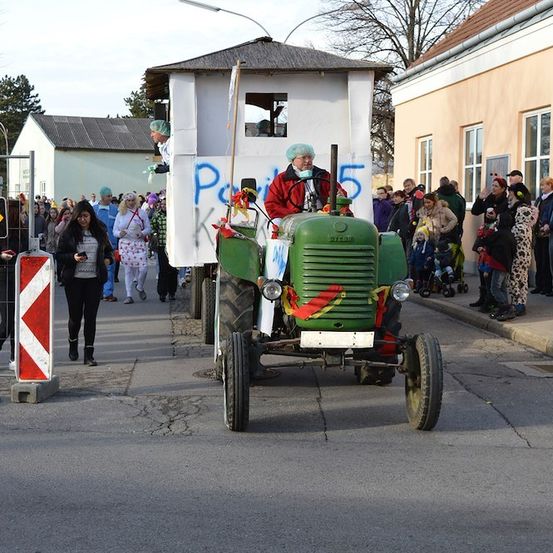 Ein grüner Traktor mit einer weißen Box auf dem Rücken fährt eine Straße hinunter, während Menschen in Kostümen zusehen.