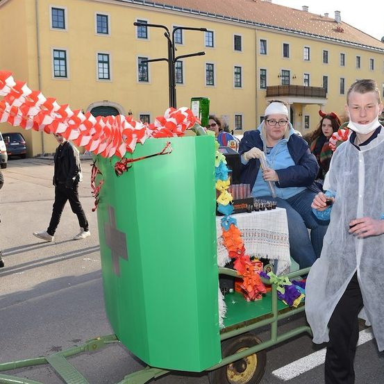 Ein Umzug mit einem grünen Wagen, der mit roten und weißen Flaggen dekoriert ist, einer Frau in einem blauen Mantel und einer Brille und einem Mann in einem Schutzanzug. Dahinter ein Gebäude mit vielen Fenstern und ein geparktes Auto davor.