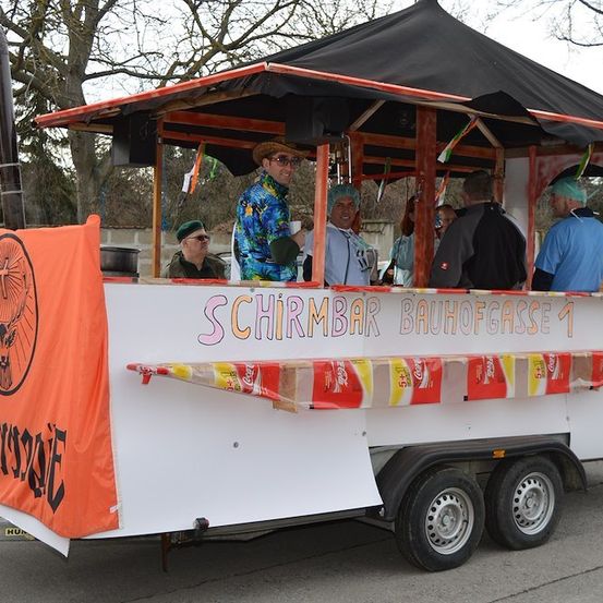 Ein bunter Anhänger mit einem Baldachin und Menschen im Inneren, er hat ein Banner mit dem Text 'SCHRMBAR BAUHOFFASE 1' und Coca-Cola-Dosen an der Seite.