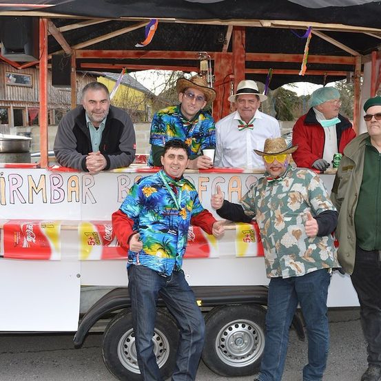 Eine Gruppe von Männern in bunten Hemden und Hüten steht vor einem dekorierten Imbisswagen mit Coca-Cola-Bannern. Sie lächeln und posieren für ein Foto.