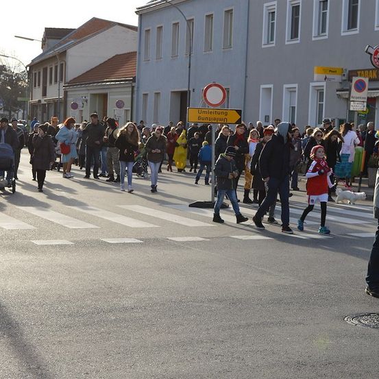 Eine Menschenmenge in bunten Kostümen überquert die Straße vor einem Gebäude mit einem gelben Schild, das 'Umleitung Umleitung' besagt.