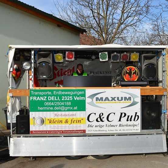 Ein mobiler DJ-Aufbau mit Lautsprechern, einem Laptop und einem bunten Papagei auf einem Anhänger. Der Anhänger hat ein Schild für Franz Deli und ein C&C Pub-Banner.
