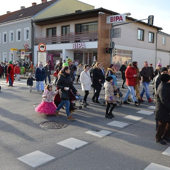 Eine Menschenmenge, einschließlich Kinder, geht über eine Straße mit Fußgängerstreifen. Einige tragen Winterkleidung und Mützen. Ein Gebäude mit einem Schild, das 'BIPA' liest, ist im Hintergrund sichtbar.