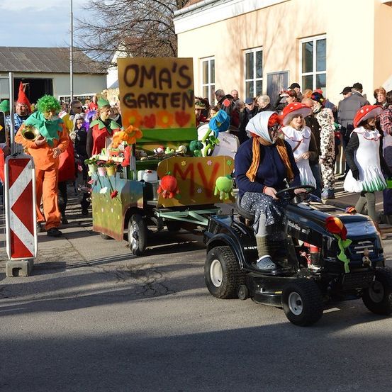 Ein Umzug mit Menschen in Kostümen und einem Wagen mit einem Schild, das Omas Garten besagt. Eine Frau fährt einen Traktor mit Anhänger.