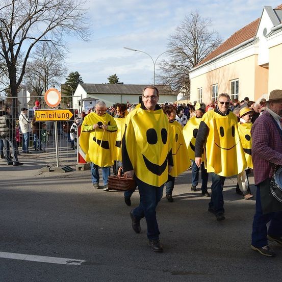 Eine Gruppe von Menschen in gelben Kostümen mit Smiley-Gesichtern geht während eines Umzugs die Straße entlang. Dahinter beobachtet eine Menge von einem Zaun aus.