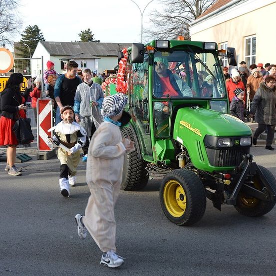 Ein Mann fährt einen grünen Traktor in einer Parade, mit Menschen in Kostümen, die um ihn herumlaufen.