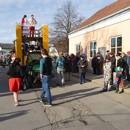 Eine Gruppe von Menschen versammelt sich vor einem Gebäude und beobachtet einen Umzug mit einem Wagen, auf dem ein Paar in traditionellen Kostümen steht. Der Wagen ist gelb und grün, und es stehen Leute auf der Straße und dem Bürgersteig.