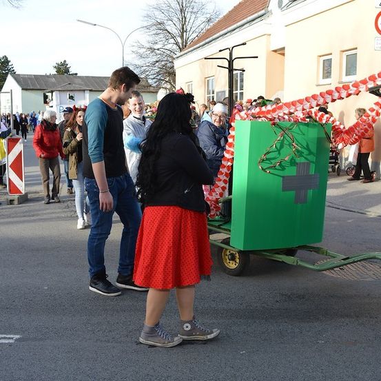 Eine Gruppe von Menschen ist um eine grüne Box mit einem Kreuz darauf versammelt, möglicherweise für eine Parade. Die Box befindet sich auf einem Anhänger mit einem rot-weißen Band. Im Hintergrund befinden sich Gebäude und Bäume.