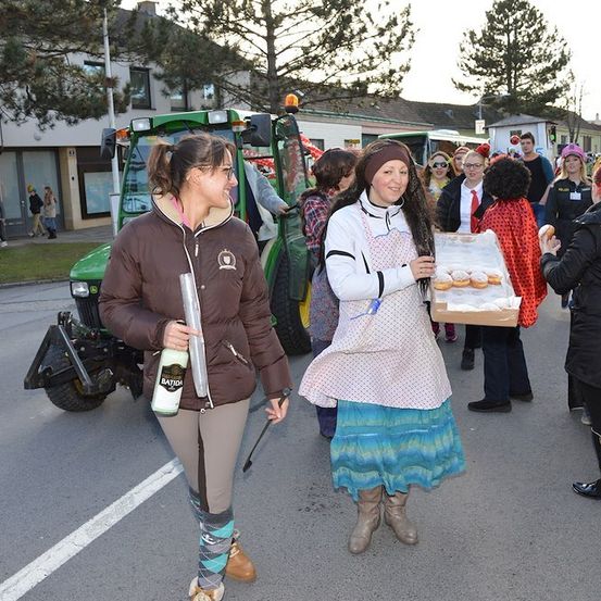 Eine Gruppe von Menschen in Kostümen geht auf der Straße. Eine Frau in einer braunen Jacke hält eine Flasche und einen Stock. Eine andere Frau hält eine Schachtel mit Donuts. Ein grüner Traktor ist am Straßenrand geparkt.