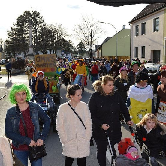 Eine lebhafte Straßenszene mit einem Umzug von Menschen in Kostümen, darunter eine Frau mit grünen Haaren, die die Straße entlanggeht. Gebäude und Bäume säumen die Straße.