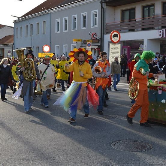 Eine Gruppe von Menschen in bunten Kostümen und Hüten zieht in einer Parade auf einer Straße. Einige spielen Musikinstrumente.