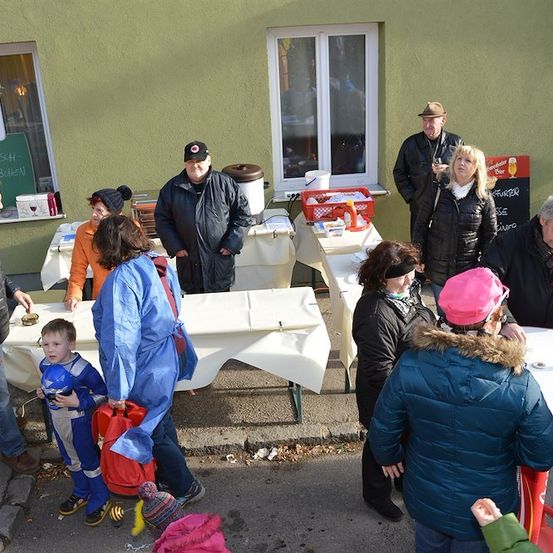 Eine Gruppe von Menschen versammelt sich vor einem Gebäude, einige stehen um Tische mit Essen und Getränken. Ein Mann mit Hut und eine Frau mit rosa Hut sind darunter. Ein Kind in einem Kostüm hält einen Helm.