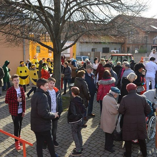 Eine Gruppe von Menschen, einige in Kostümen, versammelt sich auf einem Innenhof mit einem Baum und Gebäuden im Hintergrund.