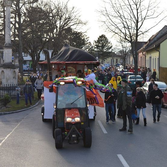 Ein Traktor mit einem dekorierten Baldachin fährt während einer Parade die Straße entlang, mit Menschen, die nebenher laufen, und geparkten Autos am Straßenrand.