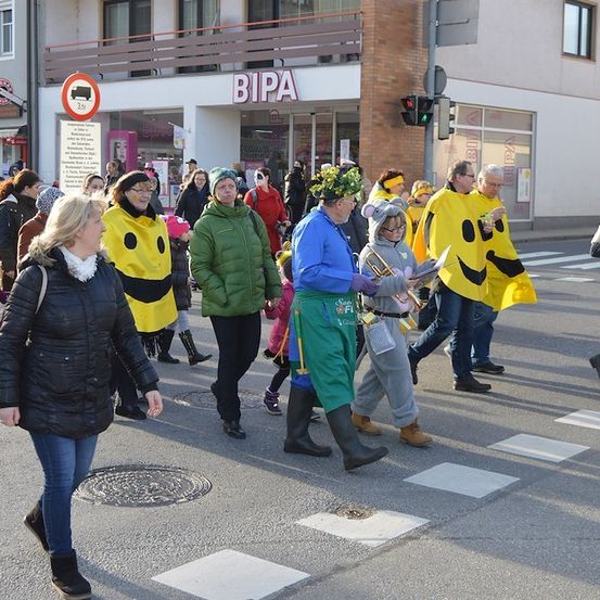 Eine Gruppe von Menschen in gelben Kostümen mit Smiley-Gesichtern zieht in einer Parade auf einer Stadtstraße. Eine Frau in einem schwarzen Mantel und Jeans geht vorneweg.