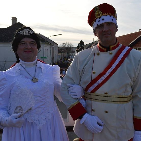 Ein Mann und eine Frau in historischen Kostümen stehen zusammen auf einem Stadtplatz, mit Gebäuden und Bäumen im Hintergrund.