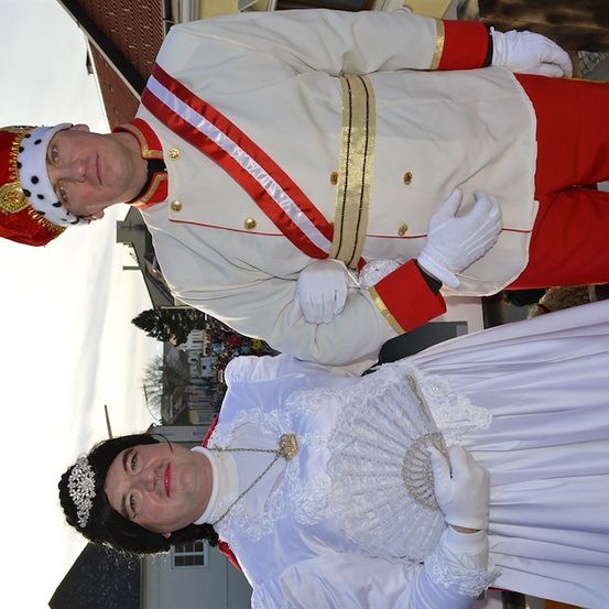 Zwei Menschen in aufwendigen Kostümen, einer in einem rot-weißen Uniform mit Krone und die andere in einem weißen Kleid mit einem Fächer, stehen zusammen vor einem Gebäude.