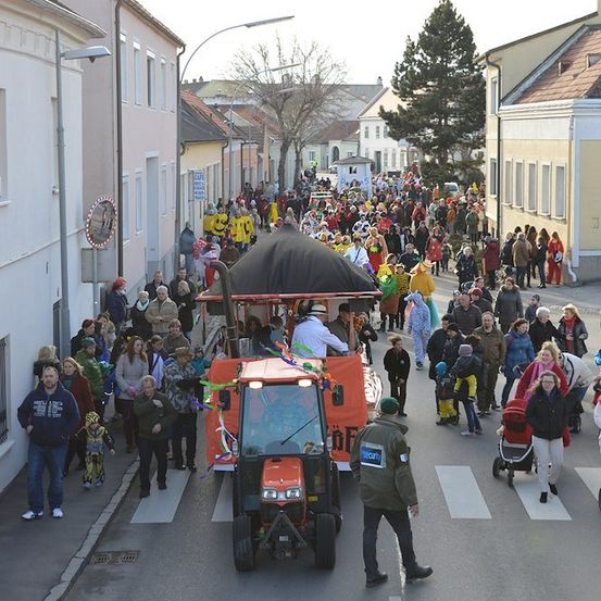 Eine große Menschenmenge in Kostümen und Hüten geht die Straße entlang, mit einem Traktor in der Mitte. Gebäude säumen die Straße, und einige Leute stehen auf dem Bürgersteig.