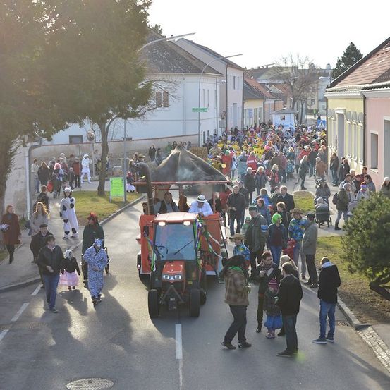 Eine große Menschenmenge in bunten Kostümen geht auf einer Straße, mit einem Traktor in der Mitte. Gebäude säumen beide Straßenseiten.