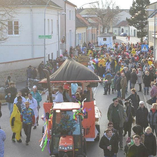 Eine Straße voller Menschen, einige in Kostümen, gehen an einem dekorierten Traktor vorbei. Gebäude säumen die Straße, und ein Schild ist im Hintergrund sichtbar.