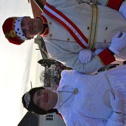 Zwei Menschen in königlicher Kleidung, einer mit Krone und der andere mit Halskette, stehen vor einem Gebäude.