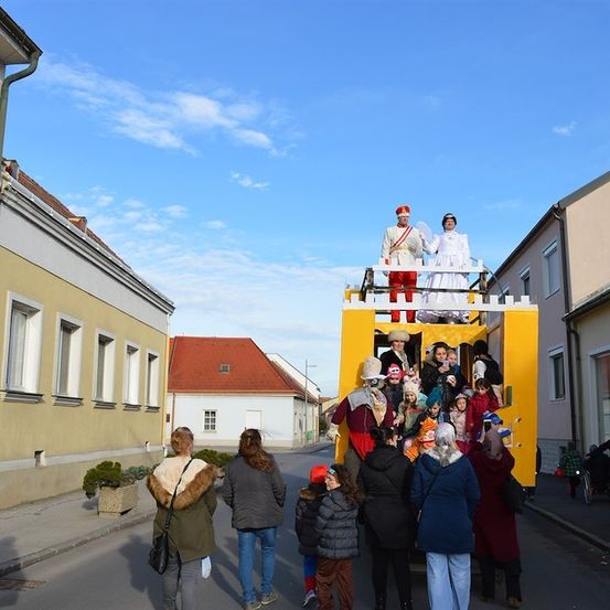 Eine Gruppe von Menschen in Kostümen fährt auf einem gelben Wagen bei einer Parade, während Zuschauer von der Straße zuschauen.