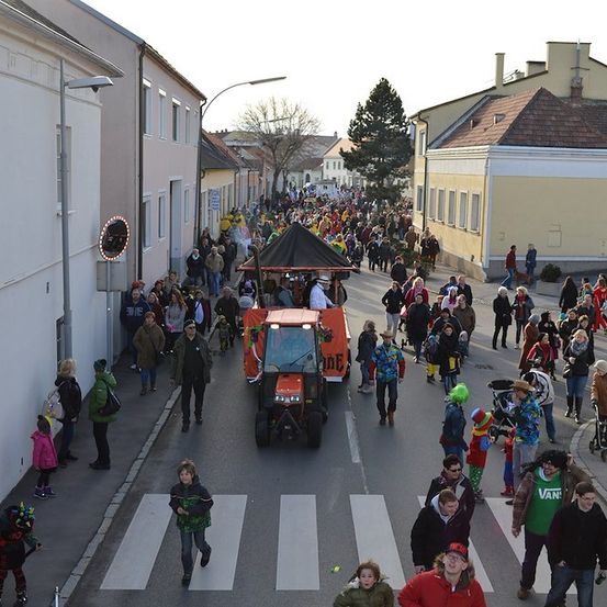 Eine große Menschenmenge, viele in Kostümen, geht eine Straße entlang, mit einem Traktor in der Mitte. Gebäude säumen beide Seiten der Straße.