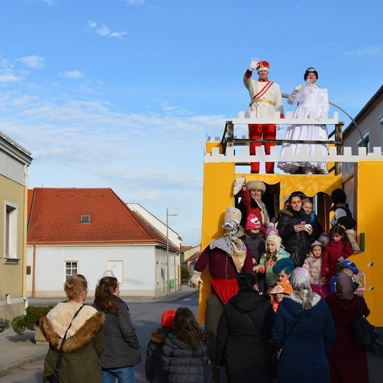 Eine Straßenszene mit einem gelben Paradewagen, der Menschen in Kostümen trägt, darunter ein Mann in einem roten Uniform, der winkt, und eine Frau in einem weißen Kleid. Zuschauer sind um den Wagen versammelt, und Gebäude sind im Hintergrund zu sehen.