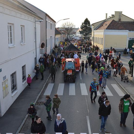 Eine große Menschenmenge, einige in Kostümen, geht auf einer Straße mit Gebäuden auf beiden Seiten, einem Traktor in der Mitte und einem Zebrastreifen.