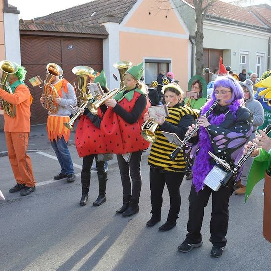 Eine Gruppe von Menschen in Kostümen spielt Musikinstrumente auf einer Straße. Sie stehen in einer Reihe und halten ihre Instrumente. Hinter ihnen befinden sich Gebäude mit Fenstern und ein Baum.