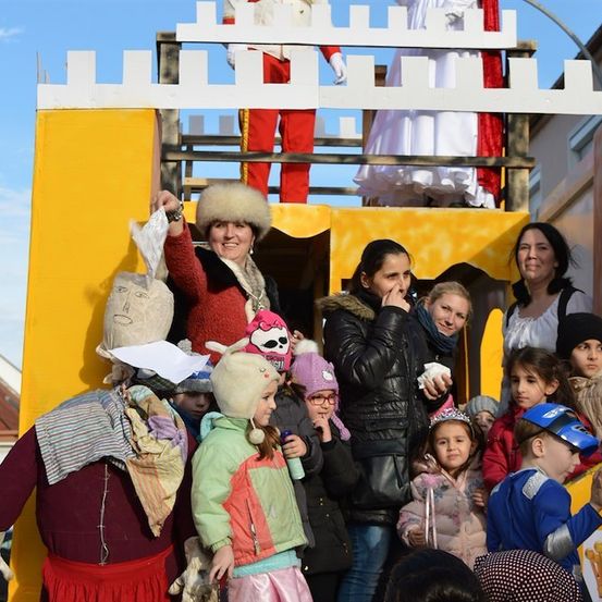Eine Gruppe von Menschen, einschließlich Kinder, versammelt sich um einen Wagen. Einige sind in Kostümen gekleidet, andere tragen Mützen und Schals. Eine Frau in einer Pelzmütze hält eine Tasche. Eine Person in roten Hosen steht auf einem Balkon.