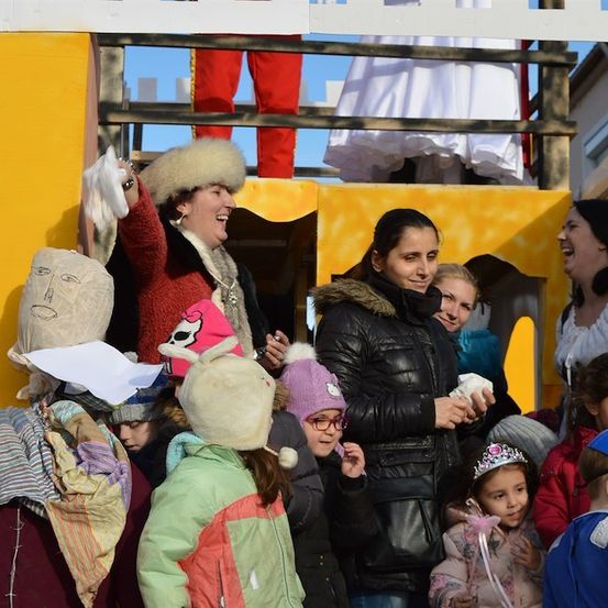 Eine Gruppe von Menschen, einschließlich Kindern, versammelt sich um einen Wagen in einem Umzug. Eine Frau in einem roten Mantel und Pelzmütze winkt der Menge zu. Ein Mädchen in einem grünen Mantel und einer rosa Mütze schaut in die Kamera.