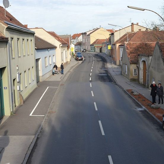 Eine ruhige Straße mit mehreren Menschen, die auf dem Bürgersteig gehen. Gebäude mit roten Dächern säumen beide Seiten der Straße. Ein Auto ist auf der Straße geparkt und ein Lieferwagen ist auf der rechten Seite geparkt.