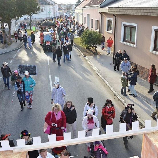 Eine große Menschenmenge in Kostümen und Hüten geht eine Straße entlang, einige tragen Kinder und Kinderwagen. Gebäude säumen die Straße, und ein Traktor ist in der Ferne zu sehen.