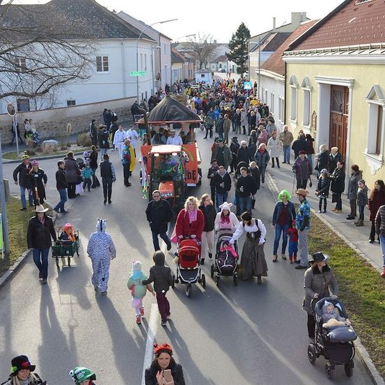 Eine große Menschenmenge, einige in Kostümen, geht eine Straße hinunter, gesäumt von Gebäuden, eines mit einem roten Dach. Ein dekoriertes Fahrzeug führt den Zug an.
