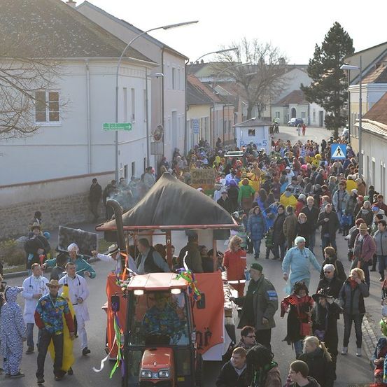 Eine große Menschenmenge in bunten Kostümen und Hüten zieht während eines Umzugs die Straße entlang. Ein Traktor mit Verdeck führt die Prozession an. Gebäude säumen die Straße, und im Hintergrund befinden sich Bäume und Straßenlaternen.