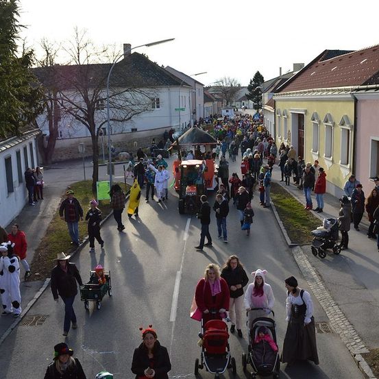 Eine große Menschenmenge in Kostümen und Hüten geht auf einer Straße in einer Stadt. Einige schieben Kinderwagen und andere tragen Kinder. Die Straße ist von Gebäuden gesäumt, einige mit roten Dächern.