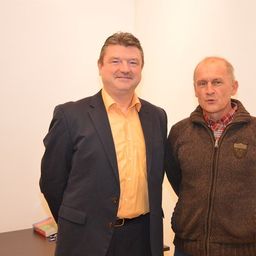 Zwei Männer stehen nebeneinander, einer in einem gelben Hemd und der andere in einer braunen Jacke, beide lächeln in die Kamera.