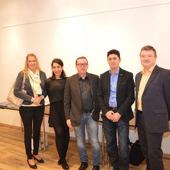 Fünf Personen, drei Männer und zwei Frauen, stehen zusammen in einem Raum mit einer weißen Wand und einem Holzboden. Sie sind formell gekleidet und scheinen für ein Foto zu posieren.