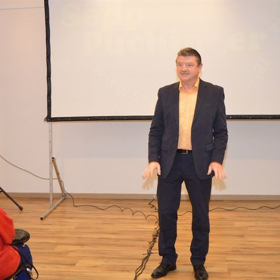 Ein Mann in einem gelben Hemd und einem marineblauen Blazer steht vor einer Projektionsleinwand in einem Konferenzraum und gestikuliert mit den Händen.