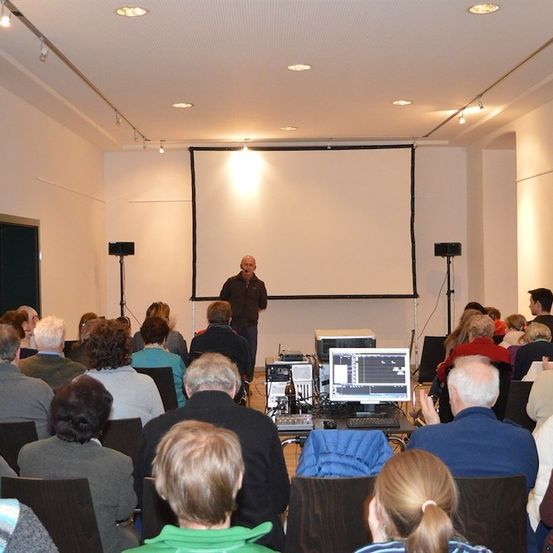Ein Mann steht an einem Rednerpult vor einer Projektionsleinwand in einem Konferenzraum und spricht eine Zuhörerschaft an.