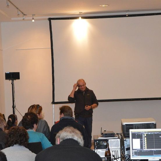 Ein Mann hält eine Präsentation in einem Konferenzraum. Er steht vor einer Projektionsleinwand mit einer leeren Folie. Vor ihm sitzen mehrere Personen.