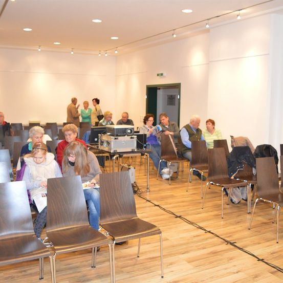 Eine Gruppe von Menschen sitzt in einem Raum auf Stühlen, einige schauen auf Papiere, während andere in Gespräche vertieft sind. Ein Beamer und ein Computer sind auf einem Tisch in der Mitte aufgestellt. Der Raum hat einen Holzboden und weiße Wände.