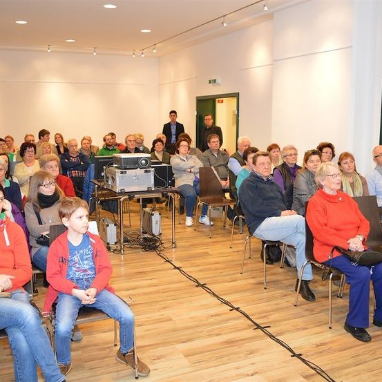 Eine Gruppe von Menschen ist in einem Raum für eine Veranstaltung versammelt. Einige sitzen, während andere im Hintergrund stehen. Geräte und Kabel sind auf dem Boden sichtbar.