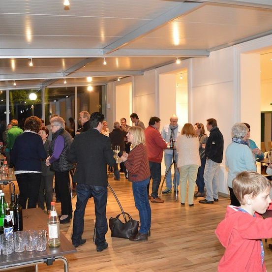 Eine Gruppe von Menschen sozialisiert in einem Raum mit Holzböden, Tischen und einer Bar-Ecke. Einige halten Gläser und Flaschen, andere stehen und unterhalten sich.
