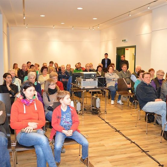 Eine Gruppe von Menschen sitzt in einem Raum, wahrscheinlich bei einer Besprechung oder Präsentation. Ein Beamer ist auf einem Tisch in der Mitte aufgestellt, und einige Personen stehen in der Nähe der Tür.