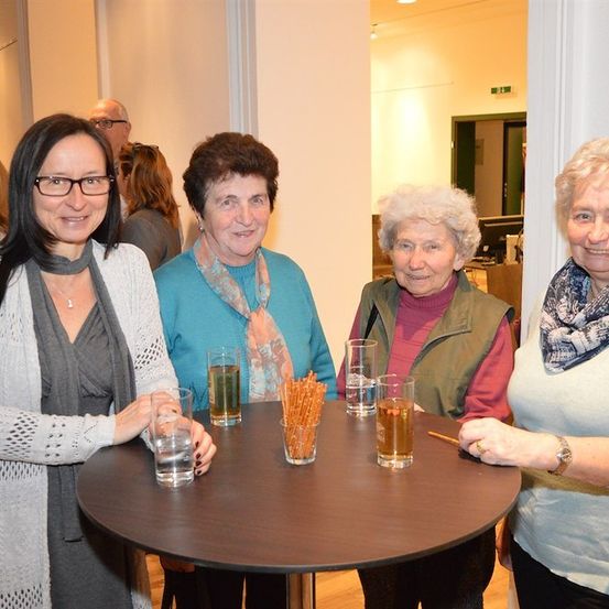 Vier Frauen lächeln und posieren für ein Foto an einem runden Tisch mit Getränken und Brezeln. Die Frau links trägt eine Brille und einen Schal. Hinter ihnen steht ein Mann in der Nähe einer Tür.