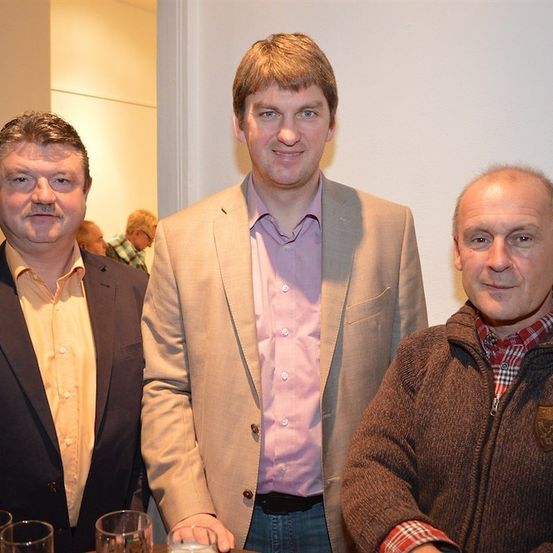 Drei Männer stehen zusammen in einem Raum und lächeln für ein Foto. Der Mann auf der linken Seite trägt einen schwarzen Anzug, der Mann in der Mitte trägt ein rosa Hemd und einen hellbraunen Blazer, und der Mann auf der rechten Seite trägt eine braune Jacke. Auf dem Tisch vor ihnen stehen Gläser und eine Kerze.